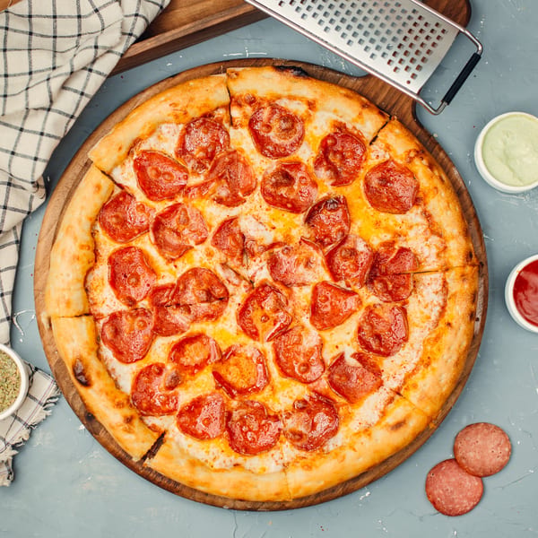 Niki's pepperoni pizza on a wooden board with sauces, a grater, and a checkered napkin.